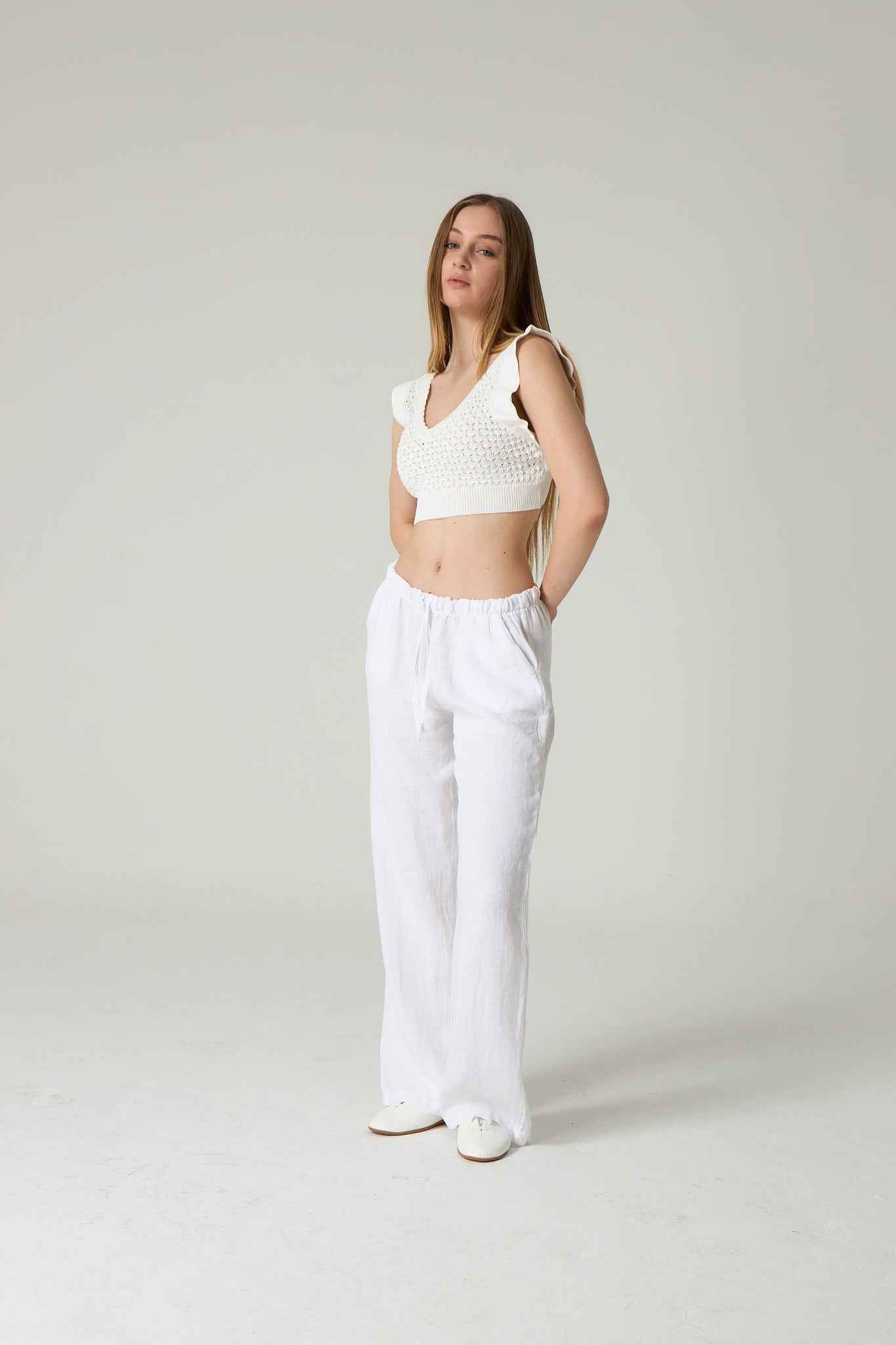 Woman wearing a white sleeveless top and white pants against a plain background.