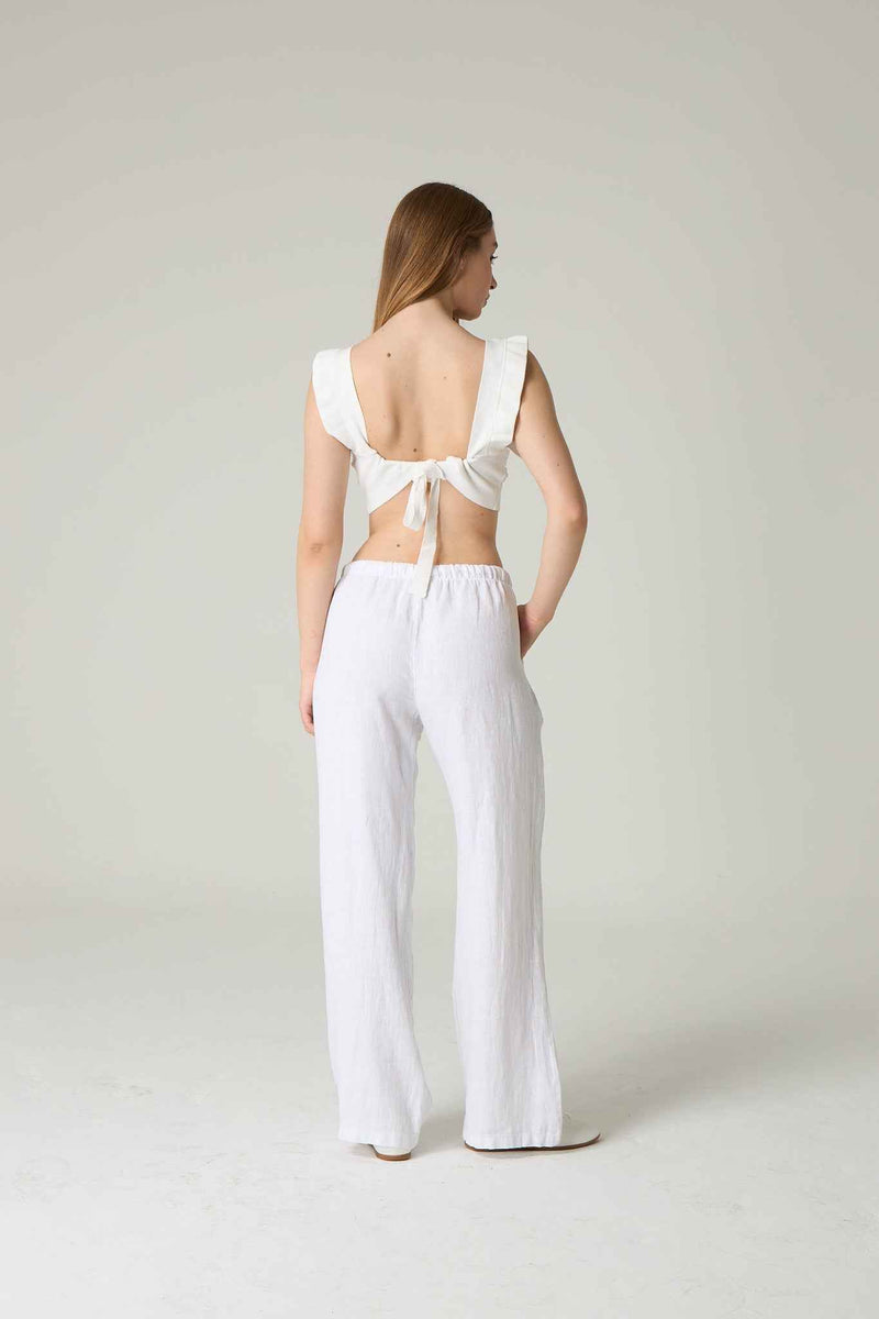 Woman wearing a white sleeveless top and pants with a bow detail on a plain background.