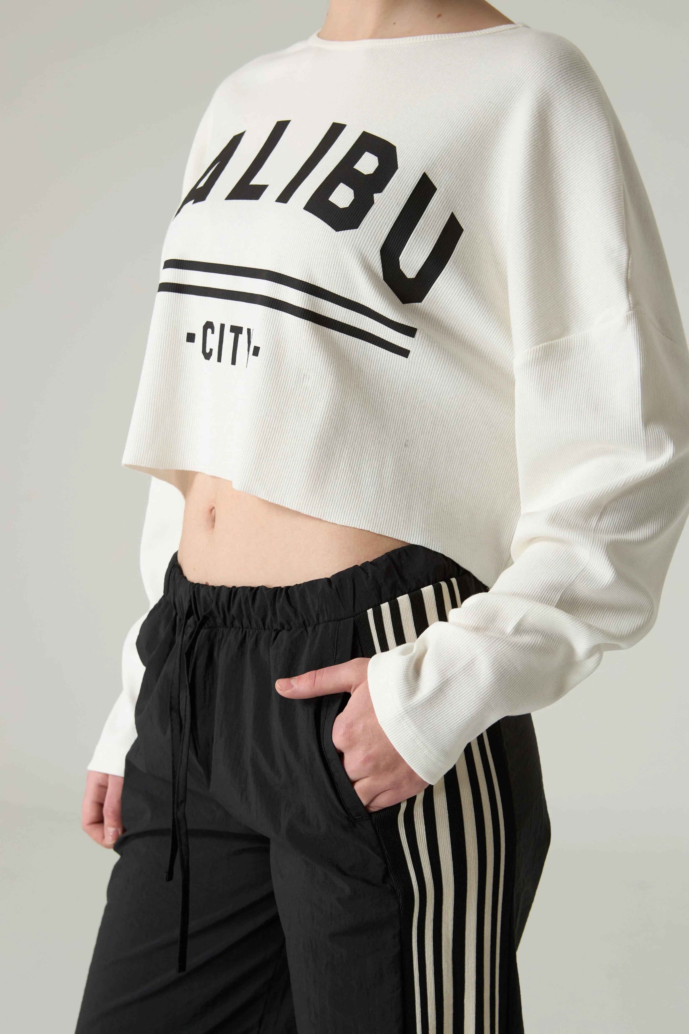 Person wearing a white cropped sweatshirt with 'MALIBU' text and black pants on a plain background.