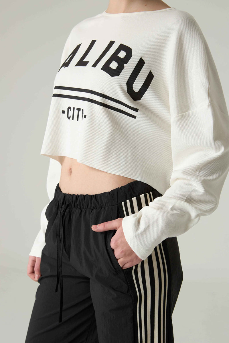 Person wearing a white cropped sweatshirt with 'MALIBU' text and black pants on a plain background.