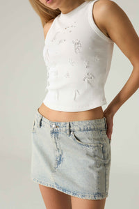 Person wearing a white sleeveless top with floral patterns and a denim skirt on a plain background.