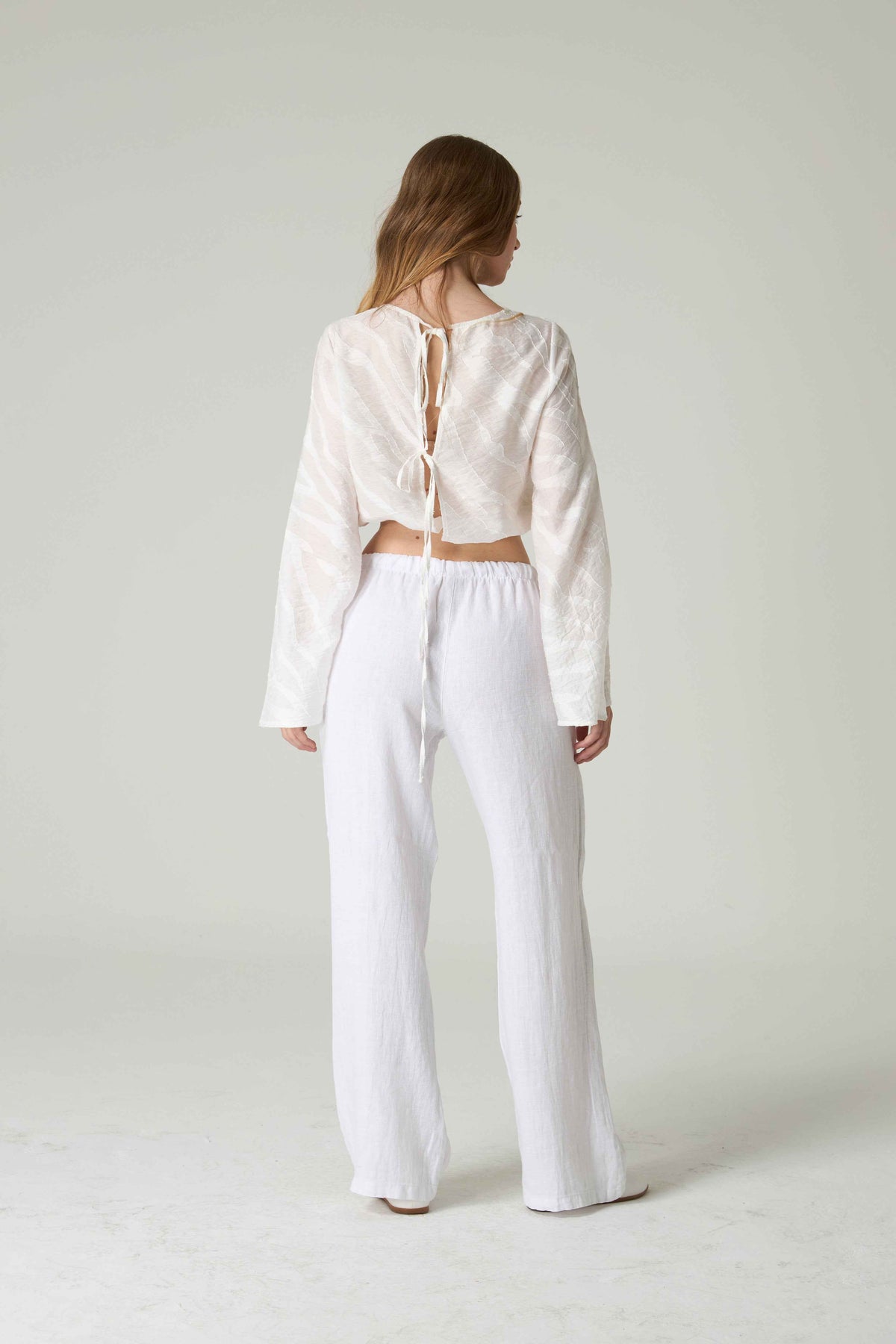 Person wearing a white lace-up top and white pants on a plain background.