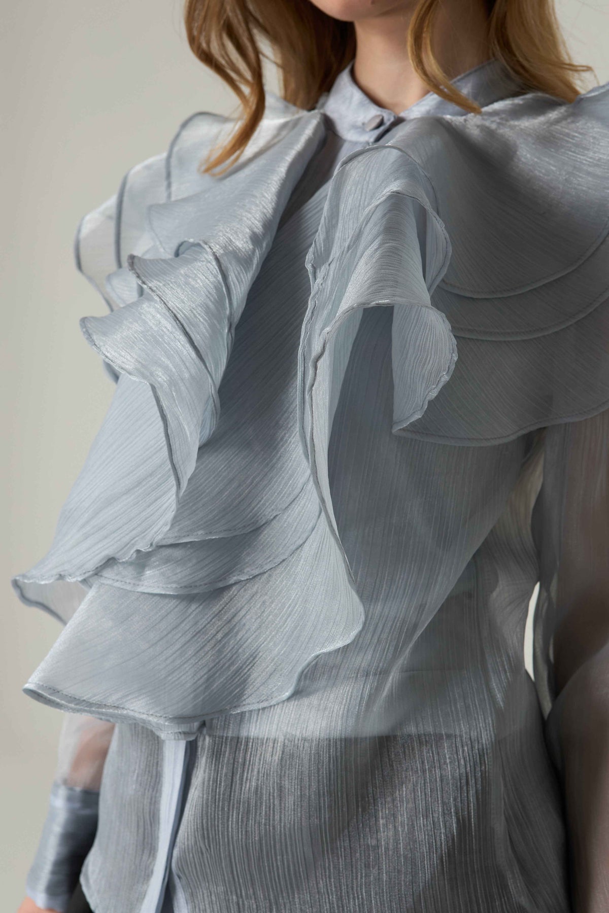 Close-up of a person wearing a light gray ruffled blouse against a neutral background.