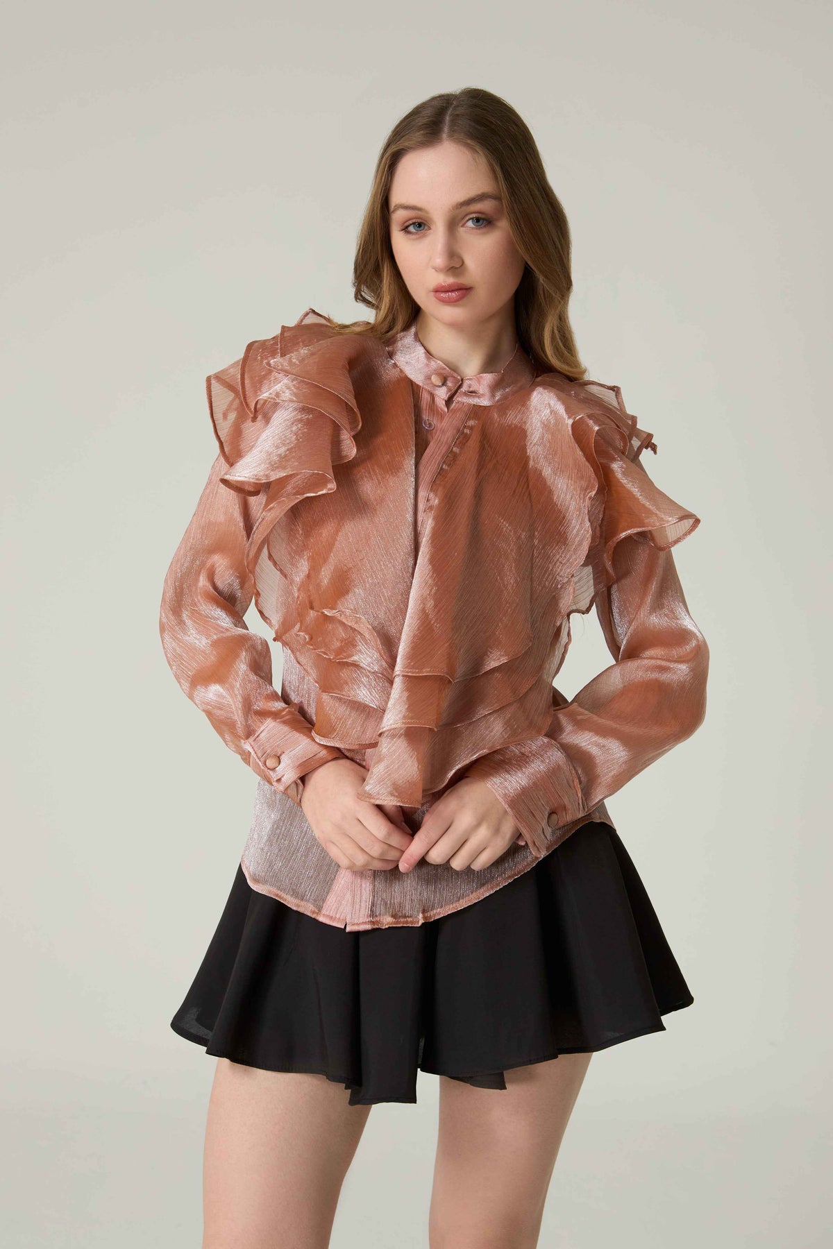 Woman wearing a sheer pink blouse with ruffles and a black skirt on a plain background.