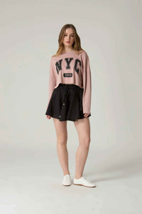 Woman wearing a pink 'NYC' sweater and black skirt on a plain background.