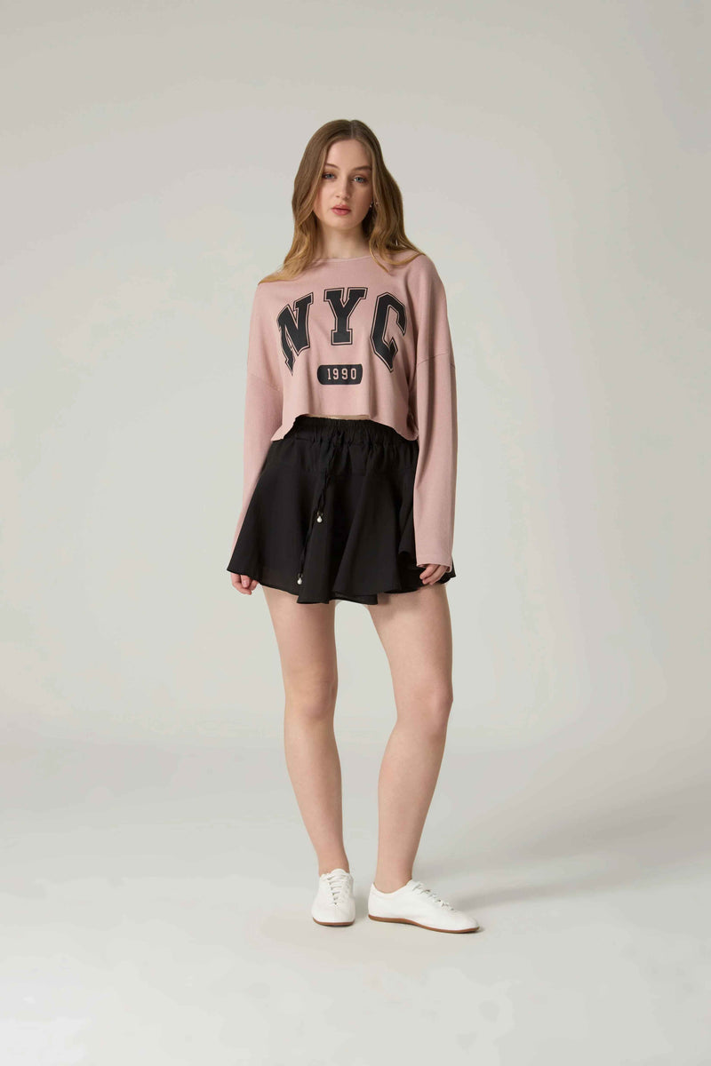 Woman wearing a pink 'NYC' sweater and black skirt on a plain background.