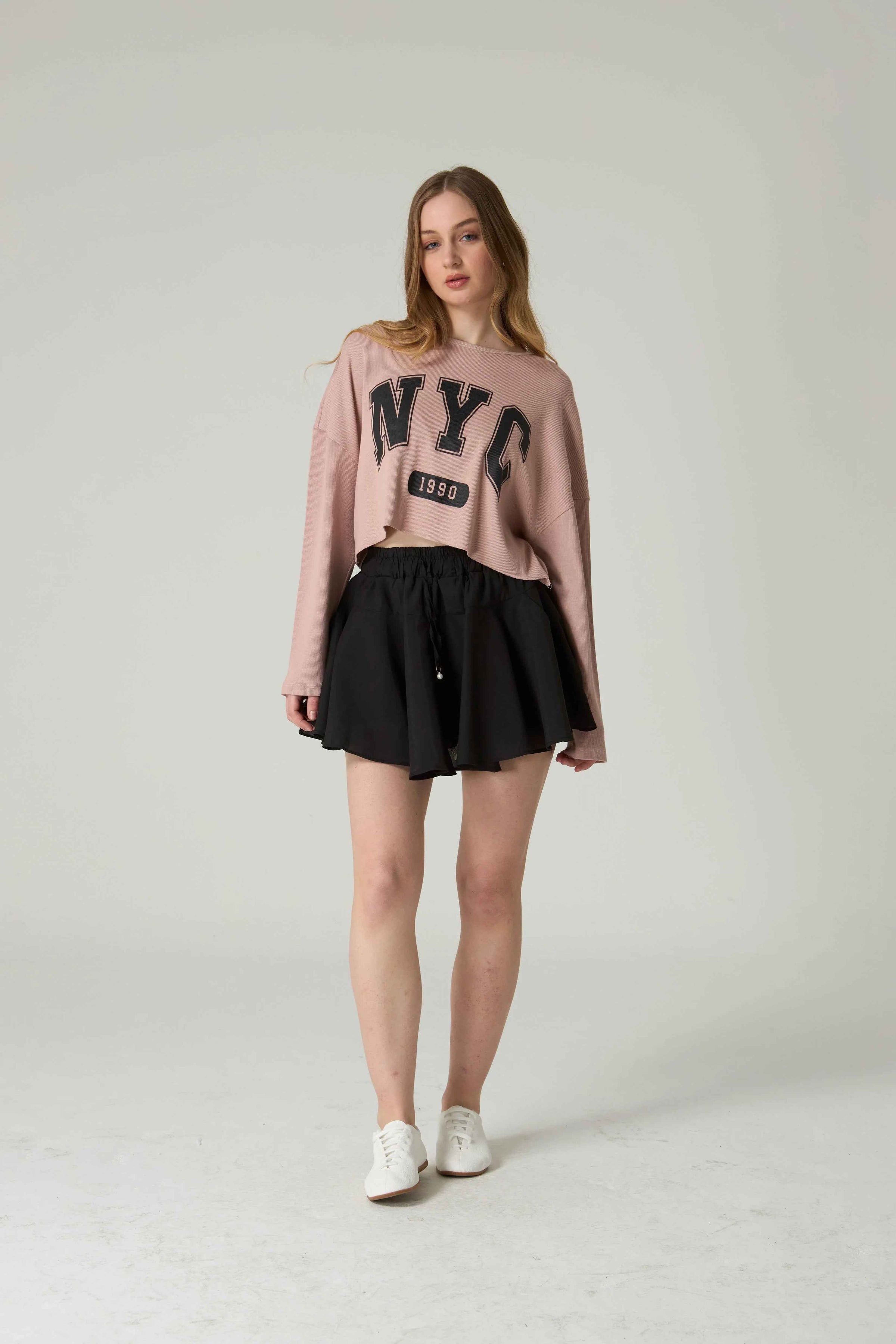 Woman wearing a pink 'NYC' sweatshirt and black skirt on a white background.