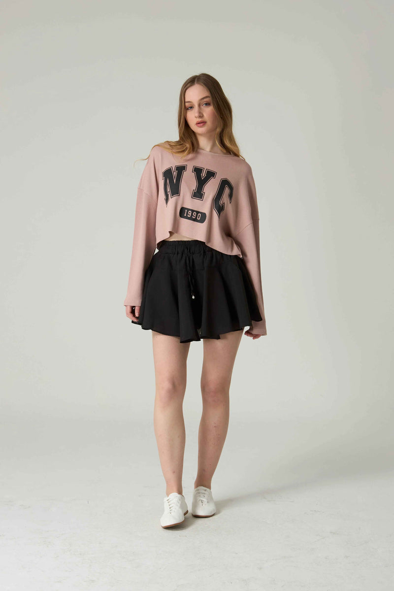 Woman wearing a pink 'NYC' sweatshirt and black skirt on a white background.