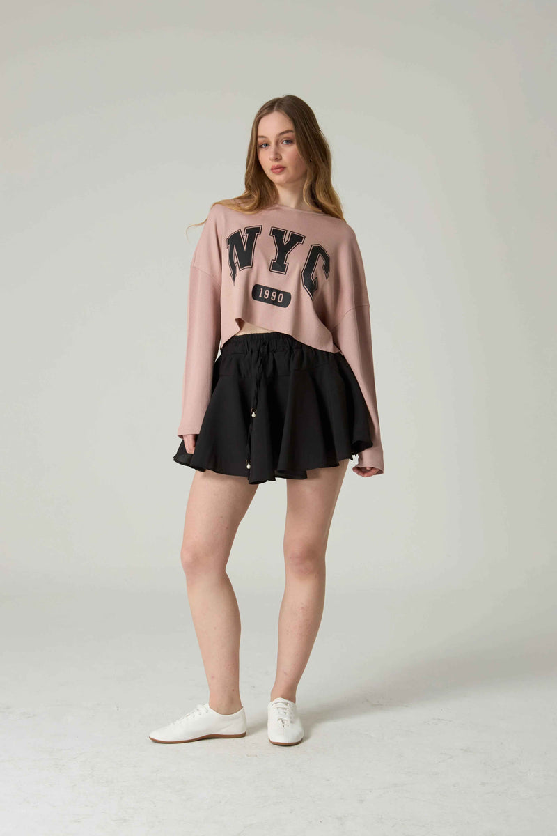 Woman wearing a pink 'NYC' sweatshirt and black skirt on a white background.
