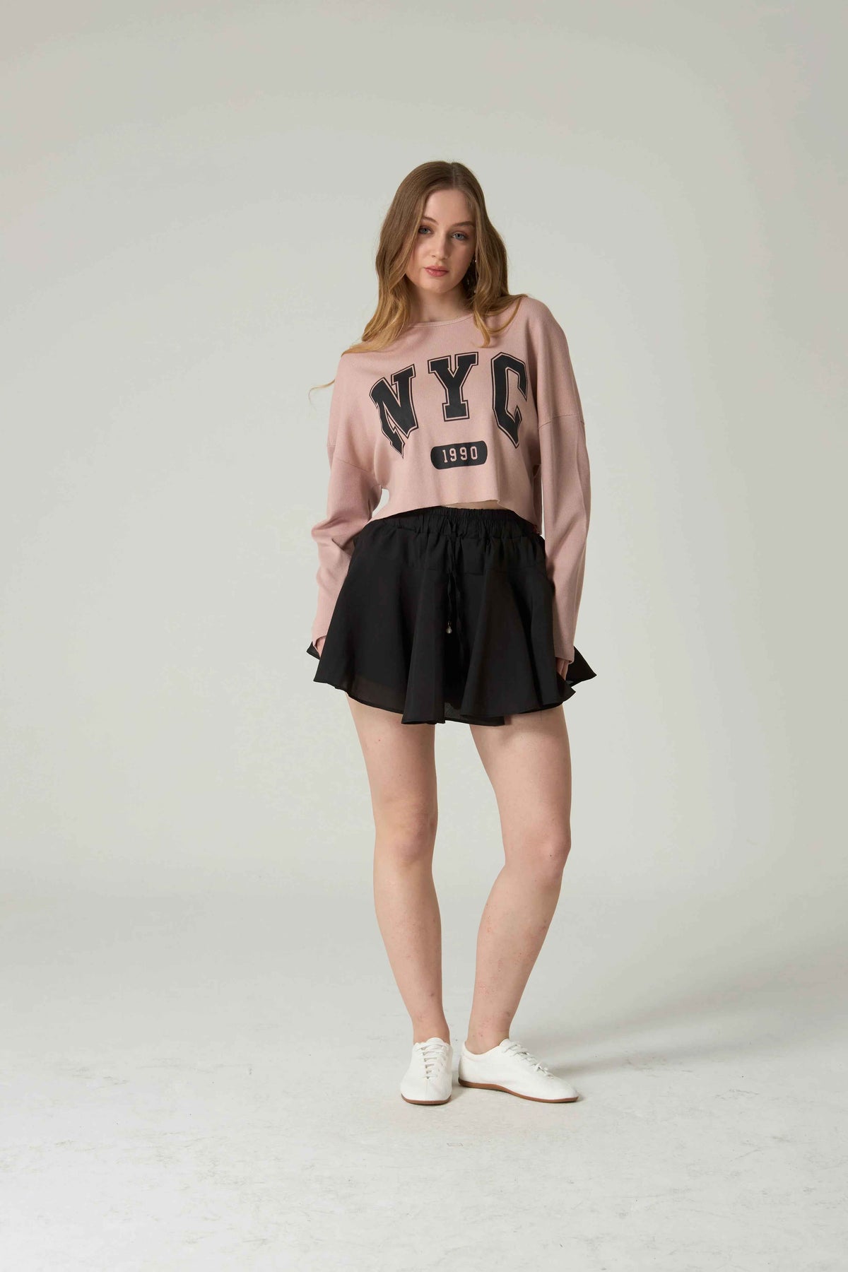 Woman wearing a pink 'NYC' sweatshirt and black skirt on a white background.