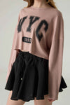 Person wearing a pink sweatshirt with 'NYC 1990' text and a black skirt on a plain background.