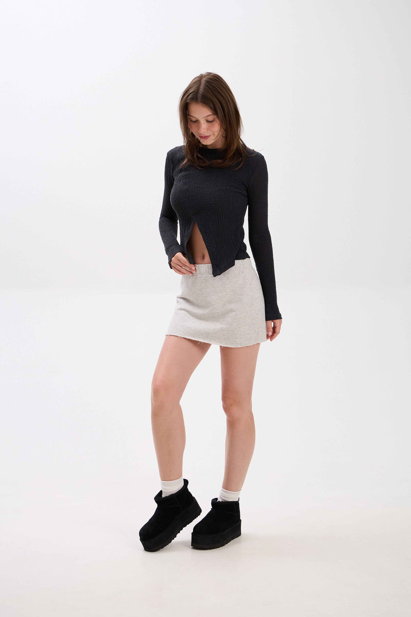 Woman wearing a black long-sleeve top with cut-out design, light gray skirt, and black shoes on a white background.