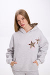 Woman wearing a light grey hoodie with leopard print star patches on the chest and a leopard print lining inside the hood, paired with matching sweatpants, standing against a plain white background.