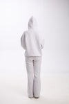 Back view of a woman wearing a light grey hoodie with the hood up and matching grey flared sweatpants, standing on a white background.