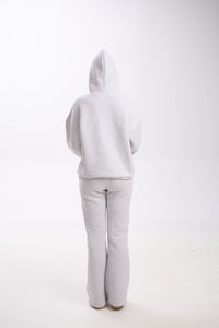 Back view of a woman wearing a light grey hoodie with the hood up and matching grey flared sweatpants, standing on a white background.