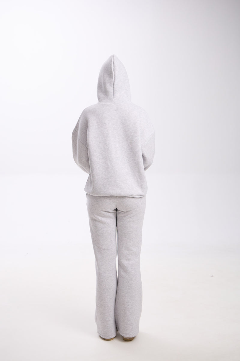 Back view of a woman wearing a light grey hoodie with the hood up and matching grey flared sweatpants, standing on a white background.