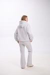 Woman standing in a light grey hoodie and matching flared sweatpants, facing slightly to the side with her back visible, against a plain white background.