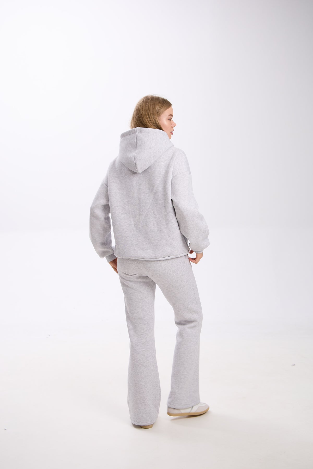 Woman standing in a light grey hoodie and matching flared sweatpants, facing slightly to the side with her back visible, against a plain white background.