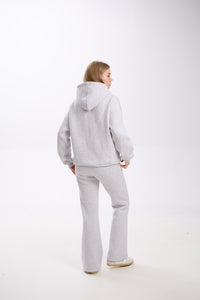 Woman standing in a light grey hoodie and matching flared sweatpants, facing slightly to the side with her back visible, against a plain white background.