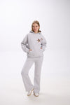 Woman posing in a light grey hoodie with two leopard print star patches on the chest, paired with matching grey flared sweatpants and white sneakers, standing against a plain white background.