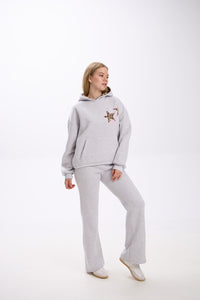 Woman wearing a light grey hoodie with two leopard print star patches on the chest and matching grey flared sweatpants, posing in white sneakers against a plain white studio background.