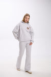 Woman posing in a light grey hoodie with two leopard print star patches on the chest, paired with matching grey flared sweatpants and white sneakers, standing against a plain white studio background.