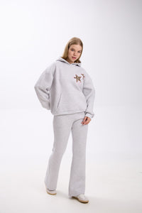 Woman posing in a light grey hoodie with two leopard print star patches on the chest, paired with matching grey flared sweatpants and white sneakers, standing against a plain white studio background.