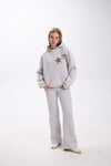 Woman wearing a light grey hoodie with two leopard print star patches on the chest, paired with matching grey flared sweatpants and white sneakers, posing with hands in pockets against a plain white studio background.