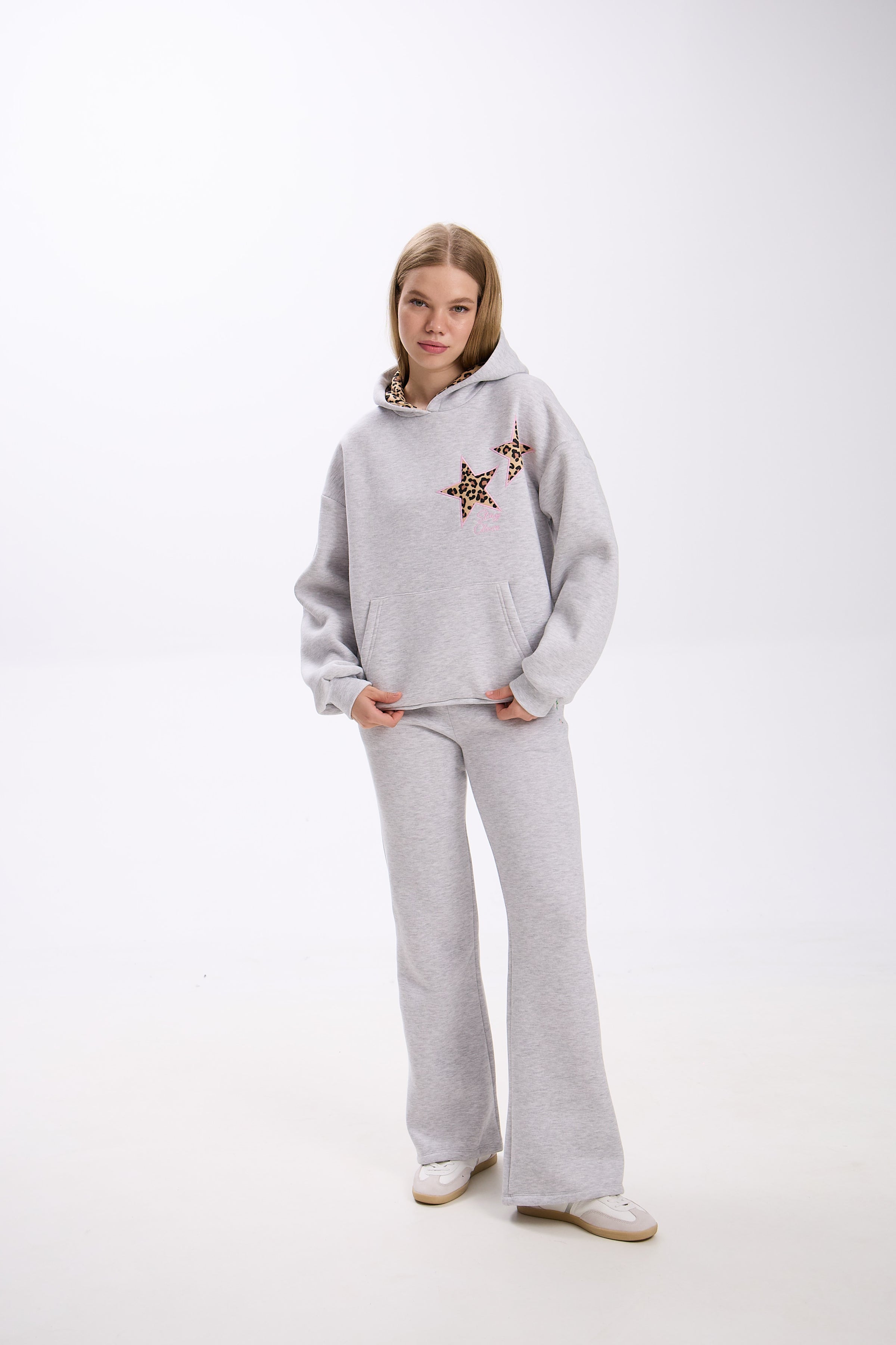 Woman wearing a light grey hoodie with two leopard print star patches on the chest, paired with matching grey flared sweatpants and white sneakers, posing with hands in pockets against a plain white studio background.