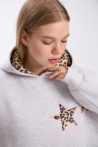 Close-up of a woman wearing a light grey hoodie with leopard print star patches on the chest and a leopard print-lined hood, gently touching her chin while looking down in a studio setting.