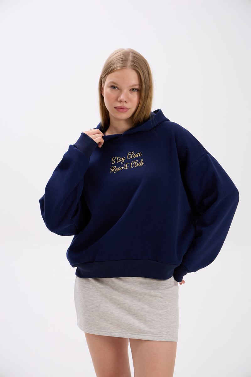 Model in a navy blue hoodie with “Stay Close Resort Club” text, paired with a light grey mini skirt.