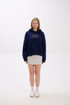 Model in a navy blue hoodie with gold “Stay Close Resort Club” embroidery, styled with a light grey mini skirt and white sneakers.