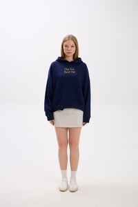 Model in a navy blue hoodie with gold “Stay Close Resort Club” embroidery, styled with a light grey mini skirt and white sneakers.