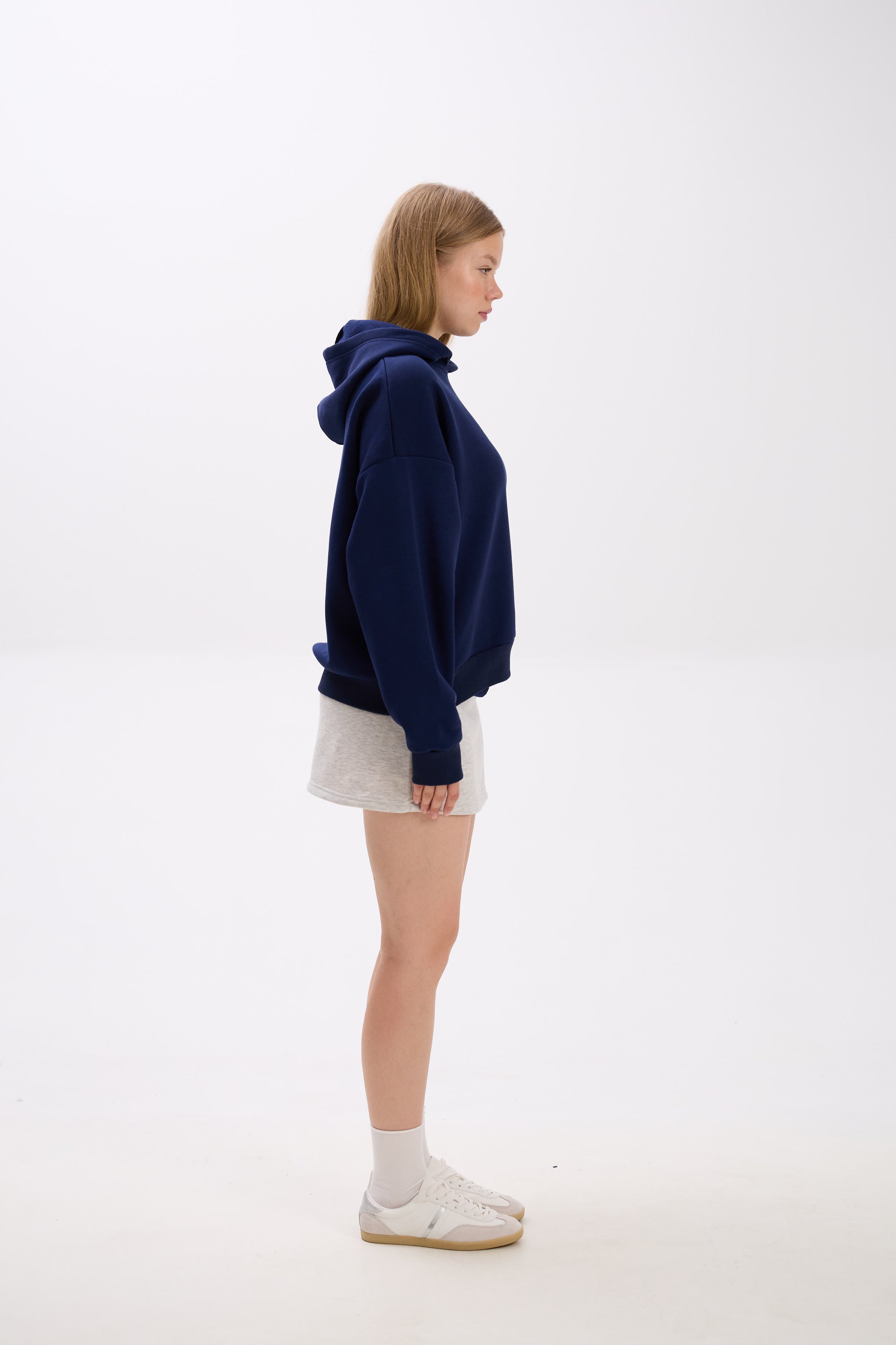 Side profile of model in a navy blue hoodie styled with a light grey mini skirt, white socks, and sneakers.