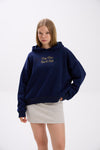 Model wearing a navy blue hoodie with gold “Stay Close Resort Club” embroidery, paired with a light grey mini skirt.