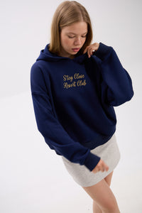 Model in navy blue hoodie with gold “Stay Close Resort Club” embroidery, paired with a light grey skirt, looking down.