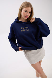 Model in navy blue hoodie with “Stay Close Resort Club” embroidery, paired with a light grey skirt, casual chic look.