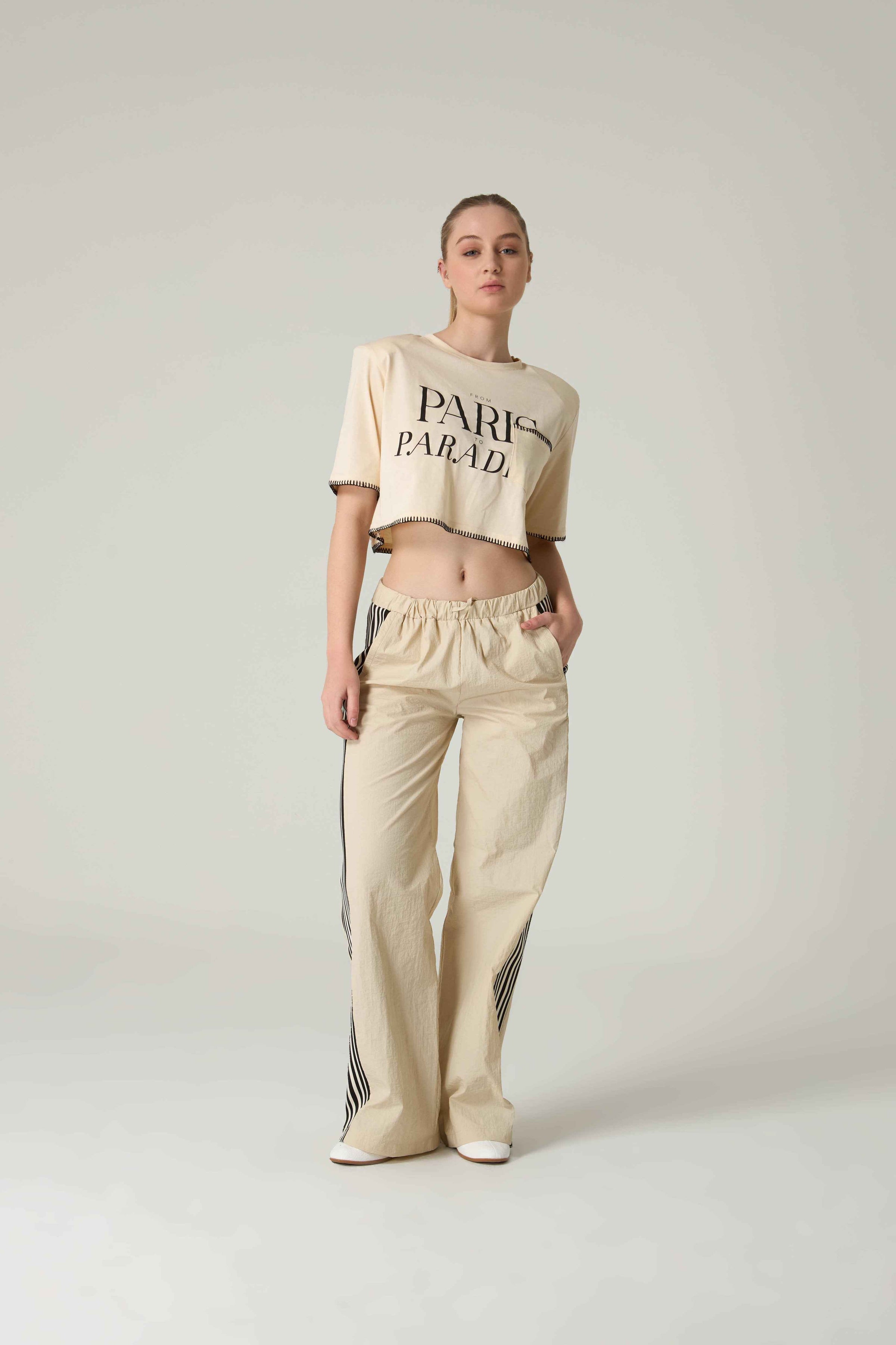 Person wearing a beige crop top with 'Paris Parade'  embroidery and pants on a light gray background.