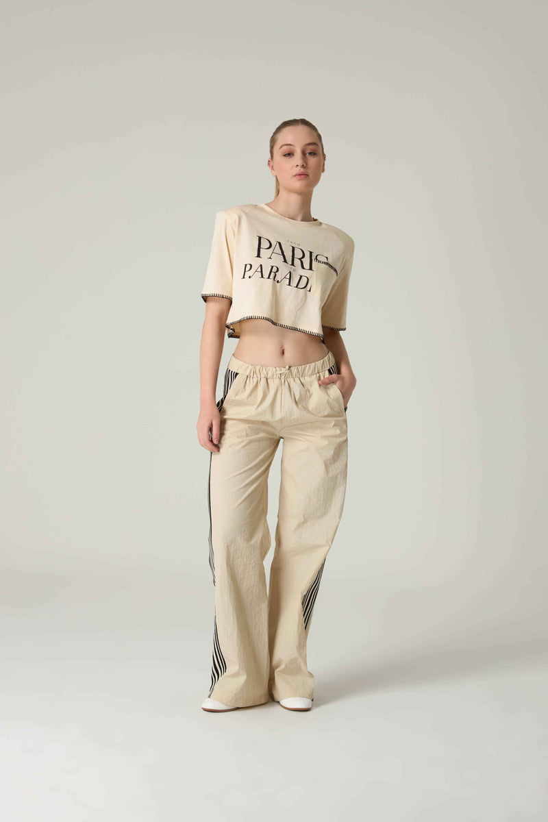 Person wearing a beige crop top with 'Paris Parade'  embroidery and pants on a light gray background.