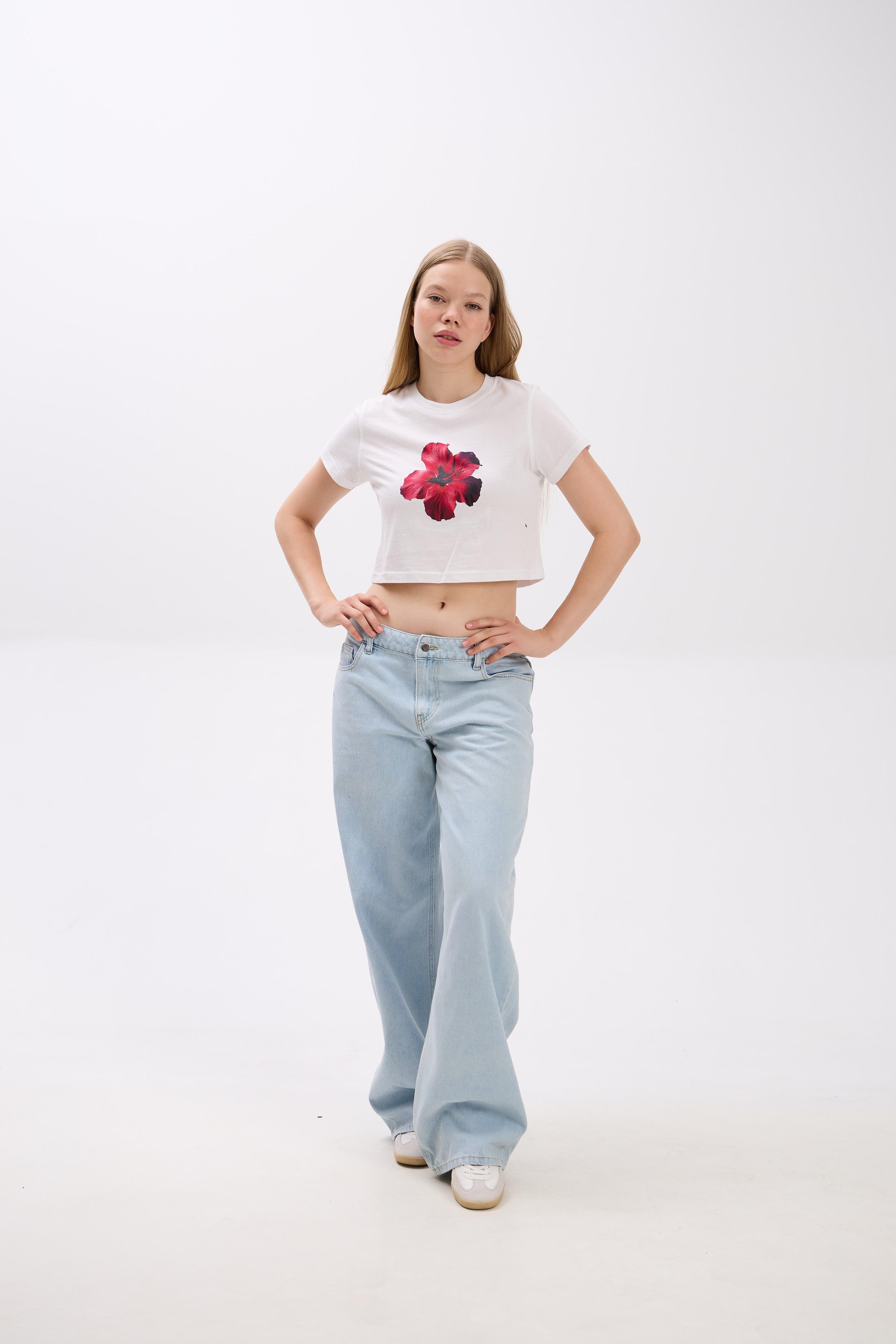 Person wearing a white crop top with a red floral design and light blue jeans on a white background.