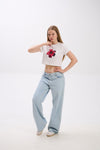 Person wearing a white crop top with a floral design and light blue jeans on a white background.