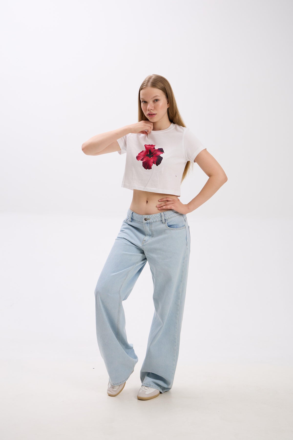 Person wearing a white crop top with a floral design and light blue jeans on a white background.