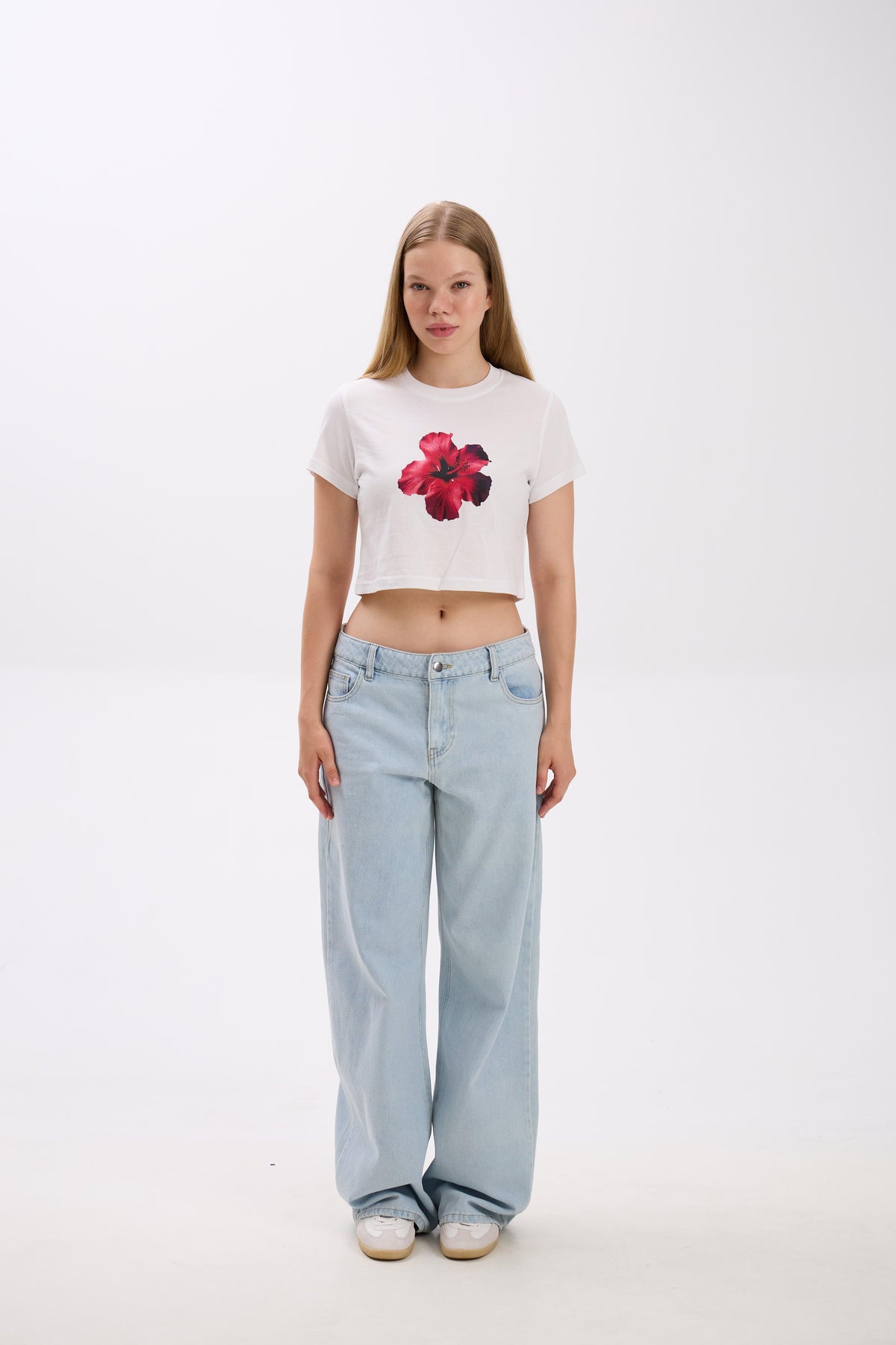 Person wearing a white crop top with a red flower design and light blue jeans on a white background.