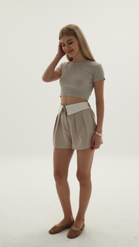 Person wearing a beige cropped short-sleeve top paired with high-waisted pleated beige shorts and brown flat shoes.