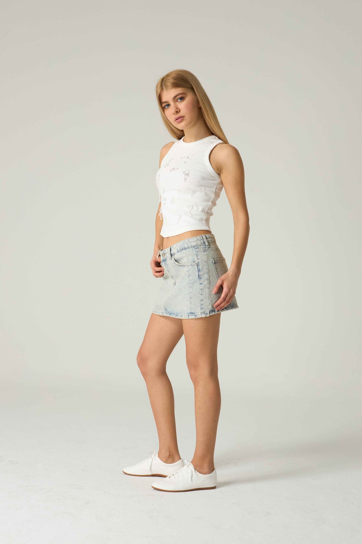 Woman wearing a white sleeveless top and denim skirt on a plain background.