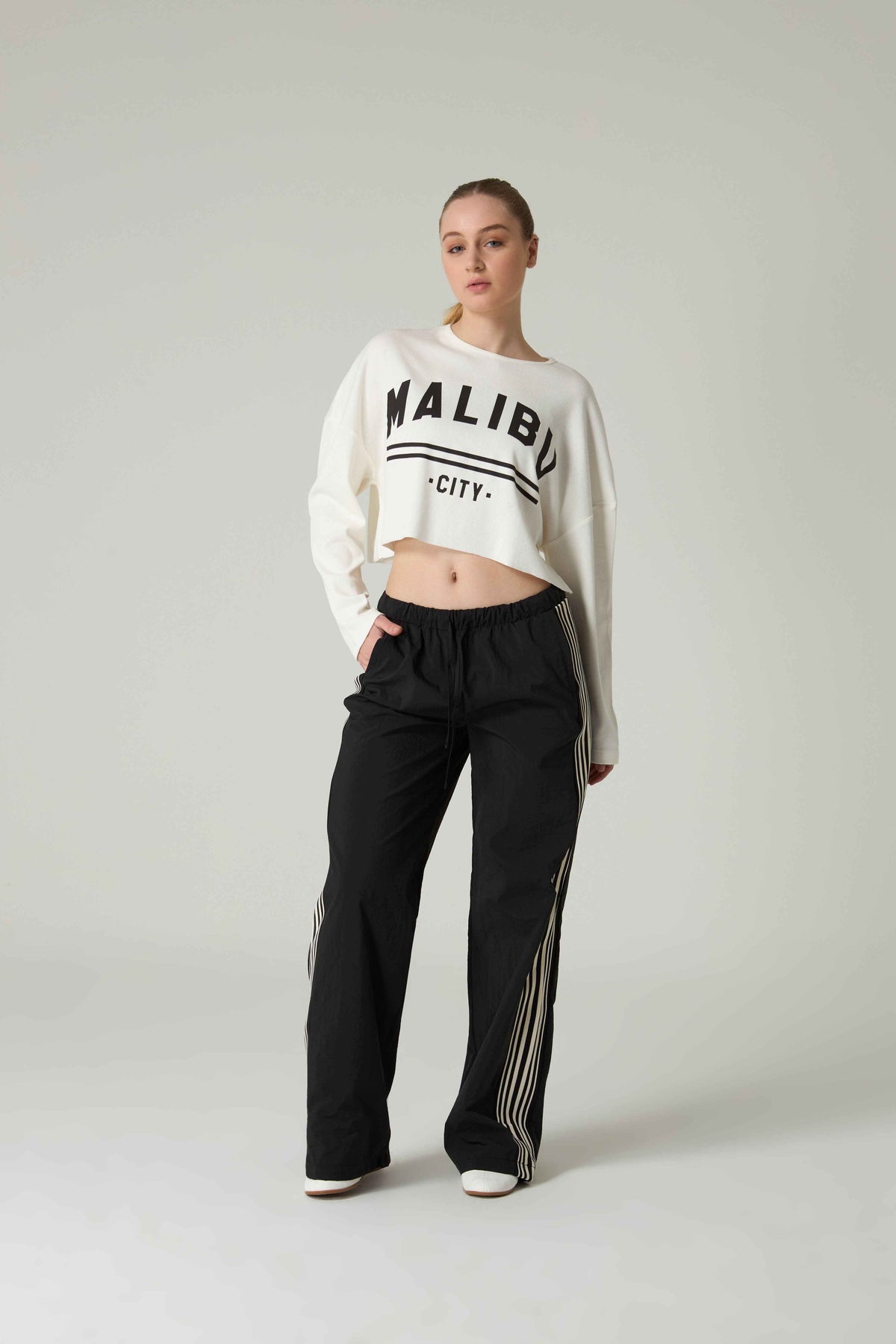 Person wearing a white sweatshirt  with 'Malibu City' embroidery and black pants on a light gray background.