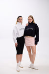 Two women wearing cropped half-zip hoodies with 'STAY CLOSE' text – one in white styled with black leggings, and the other in navy styled with a pastel pink mini skirt, both with white sneakers, against a plain background
