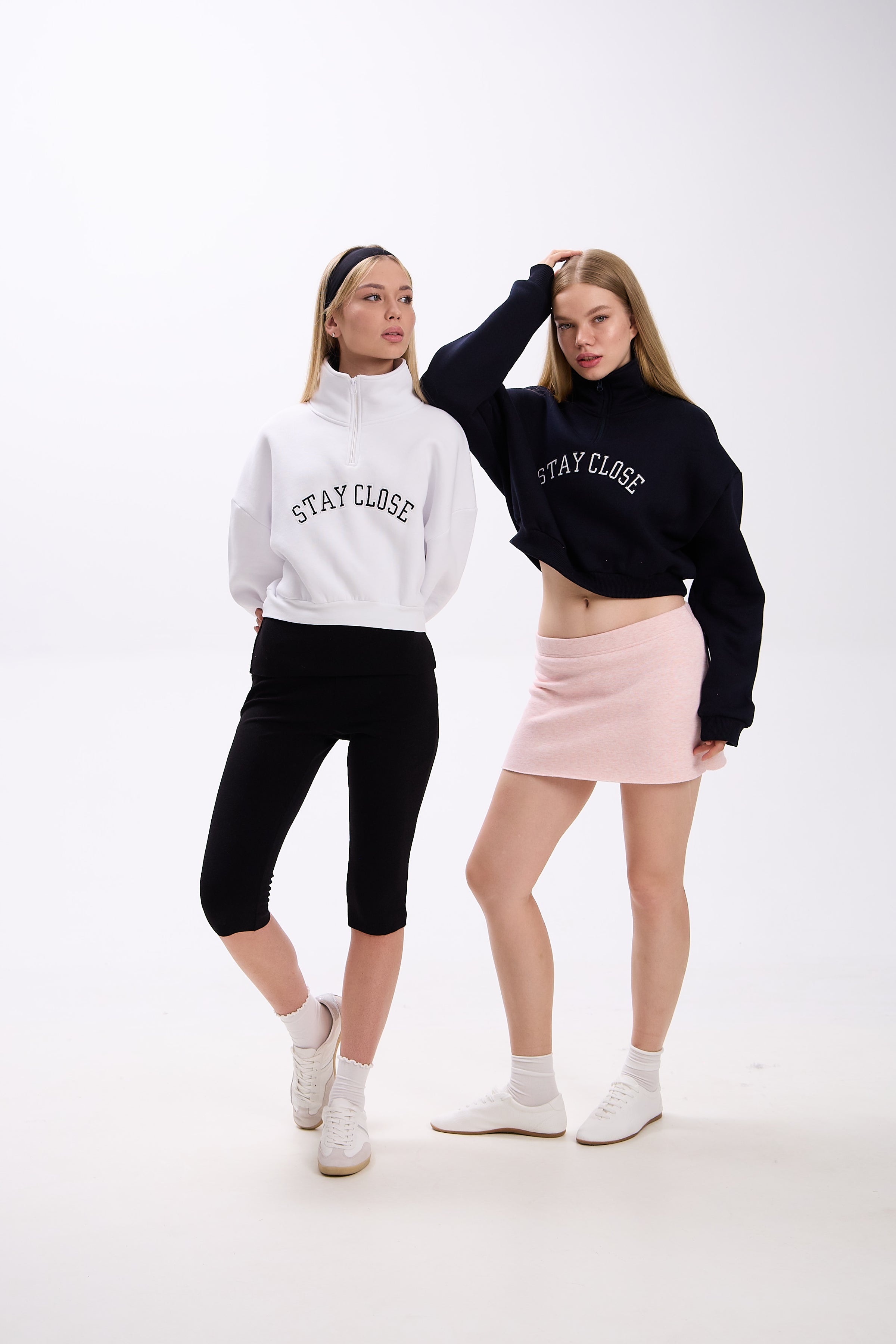 Two women wearing cropped half-zip hoodies with 'STAY CLOSE' text – one in white styled with black leggings, the other in navy styled with a pastel pink mini skirt, both in white sneakers, against a plain background