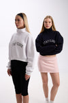 Two women wearing cropped half-zip hoodies with 'STAY CLOSE' text – one in white paired with black leggings, the other in navy paired with a pastel pink mini skirt, both styled with white sneakers, against a plain background