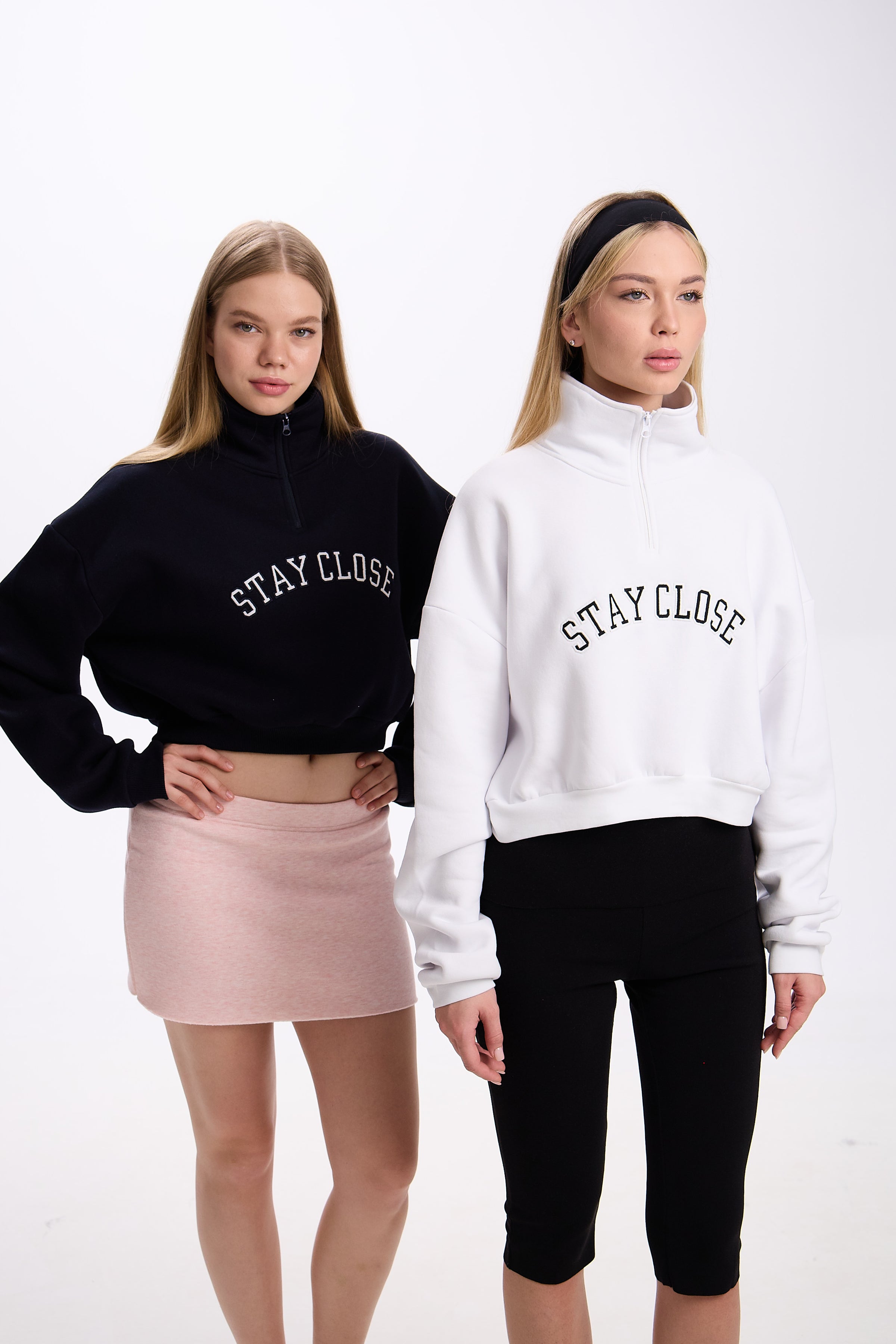 Two women wearing cropped half-zip hoodies with 'STAY CLOSE' text – one in navy styled with a pastel pink mini skirt, and the other in white styled with black leggings, both against a plain background